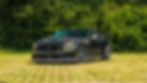 Front view of the Clive Sutton CS800DH Mustang, highlighting its aggressive carbon fibre front splitter, bonnet vents, and widebody stance, parked on a grass-paved surface with dense greenery behind.