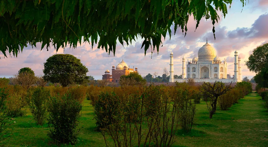 Tranquil sunset view from Mehtab Bagh, the iconic 'Moonlight Garden' offering Agra's most breathtaking perspective of the Taj Mahal across the Yamuna River. This UNESCO-protected 16th-century Mughal garden perfectly frames the Taj's reflection in its central octagonal pool, mirroring Shah Jahan's original vision of a black marble counterpart. The image captures the golden-hour glow illuminating the Taj's domes while the garden's geometric pathways, flowering shrubs, and fruit trees bloom in the foreground. A lone visitor sits on the sandstone ledge, contemplating the view where emperors once strolled. Christone Holidays' photographers reveal hidden details: the garden's alignment with the Taj's central axis, the charbagh (quadrilateral) layout symbolizing Islamic paradise gardens, and the strategic placement of cypress trees that enhance the composition. The fading light reveals the Taj's marble changing from warm gold to cool blue.
