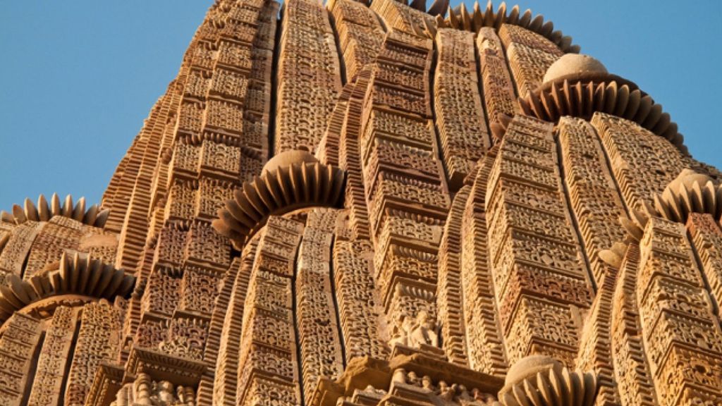 Close-up detail of Khajuraho's iconic sandstone carvings, revealing the extraordinary craftsmanship of 10th-century Chandela artisans. The weathered surface shows intricate patterns where time has softened sharp edges yet preserved astonishing details: floral motifs symbolizing fertility, geometric mandalas representing cosmic order, and narrative friezes depicting mythological scenes. The golden-hued stone bears traces of original polish in protected crevices, contrasting with wind-smoothed areas that glow under angled sunlight. Visible tool marks show how sculptors used chisels of varying widths - broader strokes for backgrounds, needle-fine points for jewelry and facial expressions. The composition highlights a transitional space where erotic Kamasutra figures blend seamlessly with divine imagery, challenging perceptions of sacred vs. sensual. Cracks and lichen growth document 1000 years of monsoon cycles.