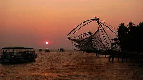 Kochi Arabian Sea Sunset Cruise Boating from Marine Drive: A mesmerizing view of a sunset cruise boat gliding across the calm Arabian Sea, departing from the iconic Marine Drive in Kochi, Kerala. The image captures the golden-orange hues of the setting sun reflecting on the shimmering water, creating a magical and romantic ambiance. The silhouette of the boat, filled with cheerful passengers, cuts through the peaceful waves with the vibrant Kochi skyline and Chinese fishing nets faintly visible in the background. The cool evening breeze ripples through the water as the sky transitions from warm tones to dusky blues. Palm-lined shores and distant ships add to the scenic charm. Marine Drive’s popular rainbow bridge and the bustling promenade are seen gently lit as twilight falls. This sunset cruise offers a tranquil and memorable experience blending city views, sea breeze, and natural beauty—perfect for travelers seeking relaxation, photography, and a glimpse of Kochi’s coastal allure.