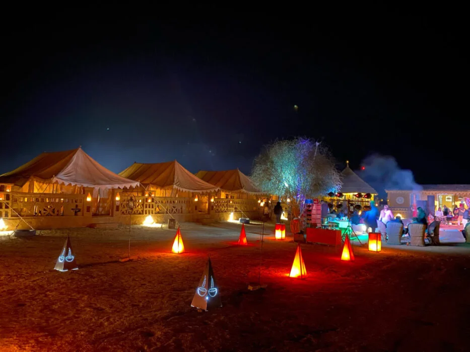 Evening spectacle at Christone Holidays' luxury desert camp near Jaisalmer, where glowing lanterns and fairy lights illuminate traditional tents against a starry Thar Desert sky. Rajasthani folk musicians perform soulful Manganiyar songs around a crackling bonfire, as guests enjoy authentic cuisine under the moonlight. This magical scene captures our signature desert camping experience - blending luxury amenities with cultural immersion. Vibrant textiles, low seating areas, and smiling travelers create a perfect snapshot of Rajasthan's nightlife. Book our Jaisalmer desert camp package for unforgettable evenings of music, dance, and stargazing in golden dunes. Where desert silence meets cultural symphony