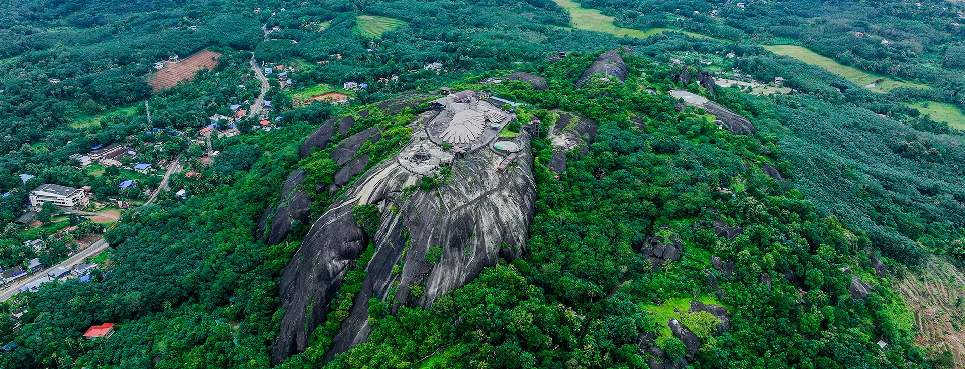A magnificent aerial view of Jatayupara, also known as Jatayu Earth's Center, located in Chadayamangalam, Kerala. The image showcases the world’s largest bird sculpture—Jatayu, a mythical eagle from the Indian epic Ramayana—perched majestically atop a rocky hill. The colossal sculpture spans 200 feet in length and stands as a tribute to the legendary bird who fought Ravana to save Sita. The surrounding landscape features vast stretches of greenery, rugged hills, and panoramic views of the Western Ghats. Visitors can be seen exploring the sculpture, adventure zones, and the cable car system that offers breathtaking rides up the hill. The sky above is bright and expansive, adding to the grandeur of the site. Jatayupara blends mythology, art, and eco-tourism, offering cultural learning, adventure activities, and scenic beauty all in one iconic destination, making it a must-visit in Kerala for both educational and recreational travel.