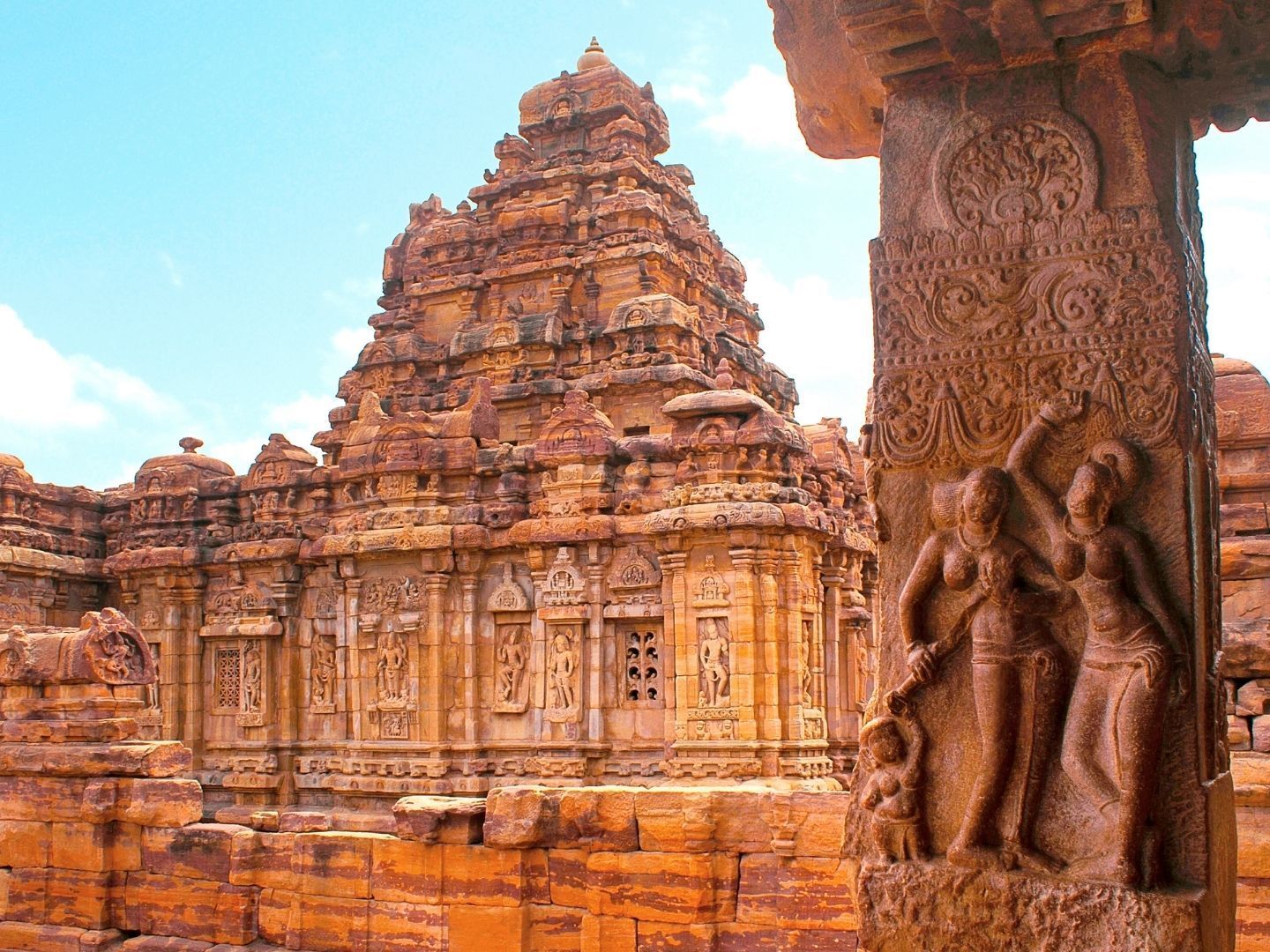 Christone Holidays presents the magnificent Sangameshwara Temple at Pattadakal - a UNESCO World Heritage Site showcasing exquisite 8th-century Chalukyan architecture. This image captures the temple's elegant Dravidian-style vimana (tower) rising majestically against Karnataka's blue skies, its sandstone walls adorned with intricate carvings of Hindu deities. The temple's square sanctum, adorned with lotus motifs, houses a sacred Shiva linga where devotees have worshipped for over 1,200 years. With Christone's knowledgeable guides, you'll discover how this temple represents the perfect fusion of North and South Indian architectural styles - notice the curvilinear tower contrasting with the tiered pyramidal roof. The surrounding complex of Pattadakal's other heritage temples creates a breathtaking panorama of India's golden age of temple construction. Christone's curated tours include special access to view the temple's remarkable carvings up close.