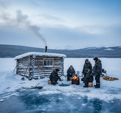 Winter survival training in Yakutia showing ice fishing through thick ice near traditional log cabin