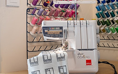 Surjeteuse Elna eXtend 664PRO en fonctionnement, cousant un tissu personnalisé "Édition Limitée Handcrafted", avec un support mural de bobines de fils multicolores en arrière-plan.