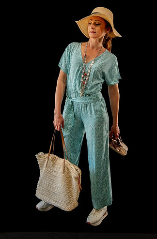 Femme de face, combinaison fluide bleu-vert à motif, chapeau de paille à large bord, sac filet écru à l’épaule et baskets blanches, sandales à la main droite.