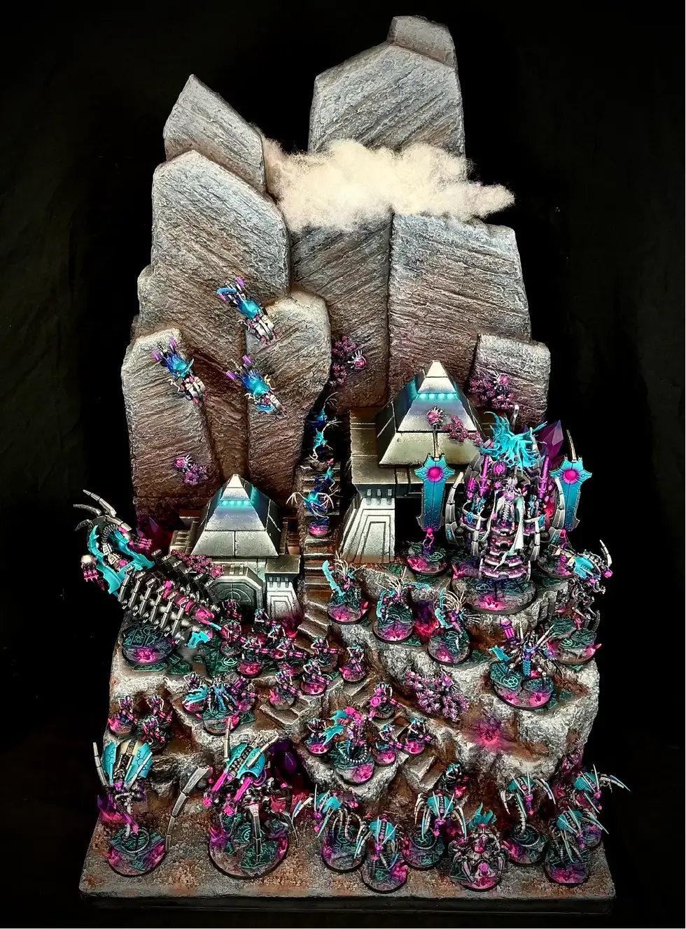 Miniature sci-fi battle scene with colorful robotic figures in pink and blue atop a rocky, mountainous terrain. Structures resemble ancient temples.