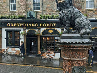 Statue of a dog on a stone pedestal in front of "Greyfriars Bobby" pub. Wet Edinburgh street, empty tables, and people walking by in coats.