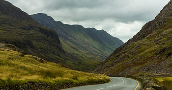 Winding road through lush green mountains beneath a cloudy sky, creating a serene and remote atmosphere.