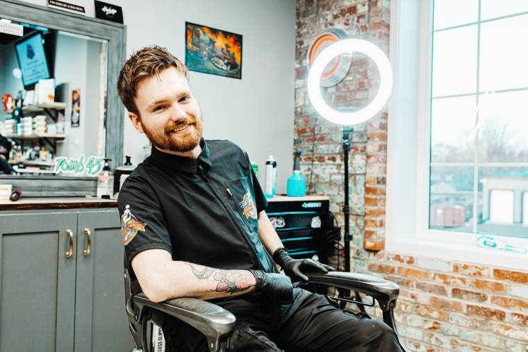 Smiling tattooed man seated in a barber chair beside a large window