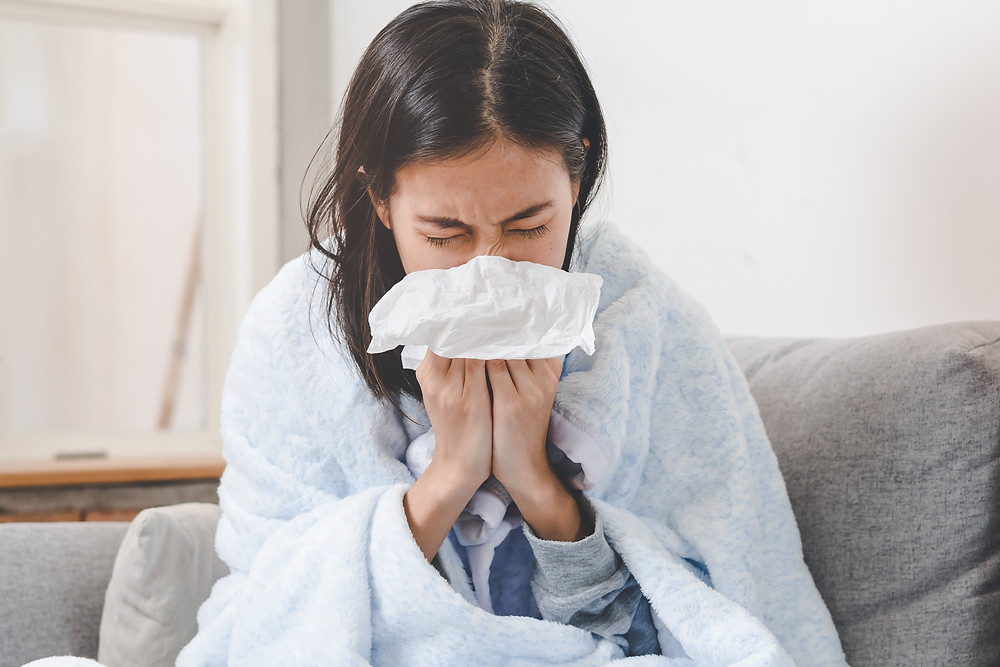 A woman sits on a couch, sneezing into a tissue, which illustrates her need to use oregano oil for a cold