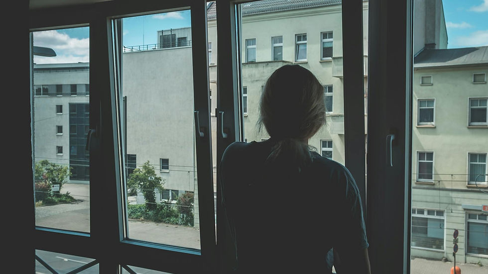 A woman stands indoors watching the cityscape through large windows, capturing a serene urban scene.