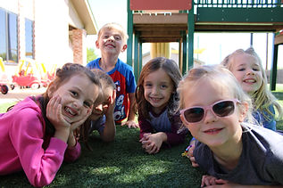 Preschool students are laying on the ground outside on a playground and smiling for the camera