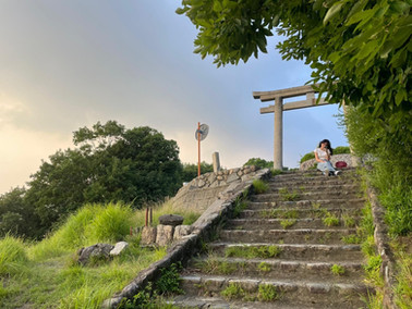 Girl sitting at the top of stone steps. Greenery visible, dusk.