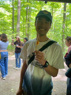 Young man smiling with maple syrup on a popsicle stick in a forest. Others in the background enjoy treats. Bright green foliage creates a cheerful mood.