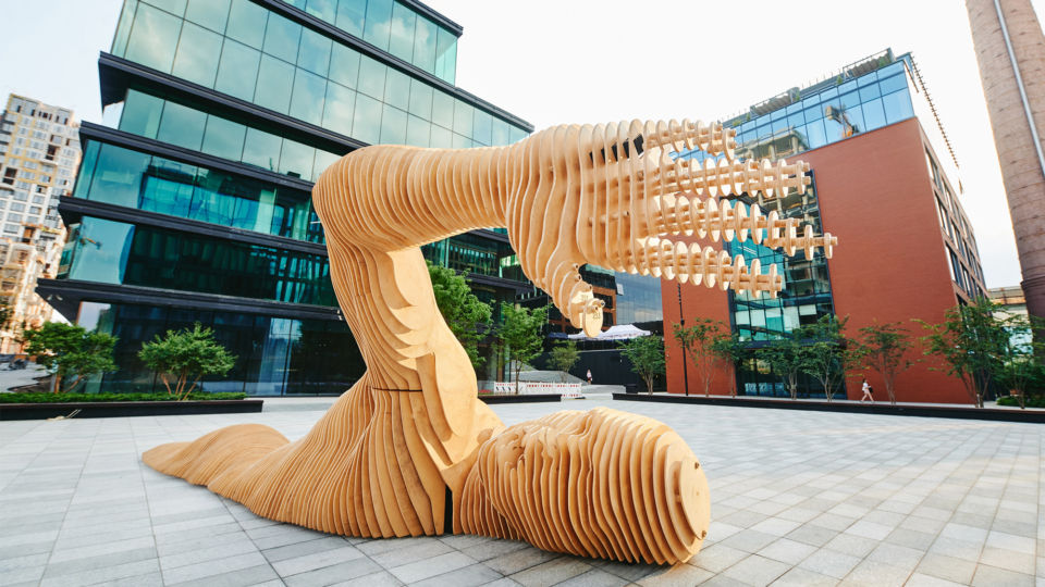 Wooden sculpture of an arm reaches upward in a modern plaza with glass buildings, trees, and a red brick wall in the background.