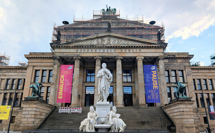 Statues in front of Berlin's Konzerthaus, scaffolding visible. Pink and blue banners with text hang from columns. Cloudy sky backdrop.