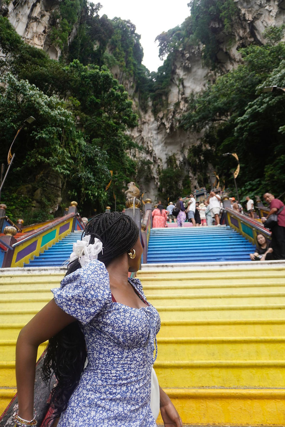 Woman in a blue dress on colorful staircase, looking back. Tourists climb steps surrounded by lush greenery and rock formations.
