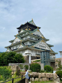 Japanese castle with man in blue shirt in the foreground. Greenery, blue skies.