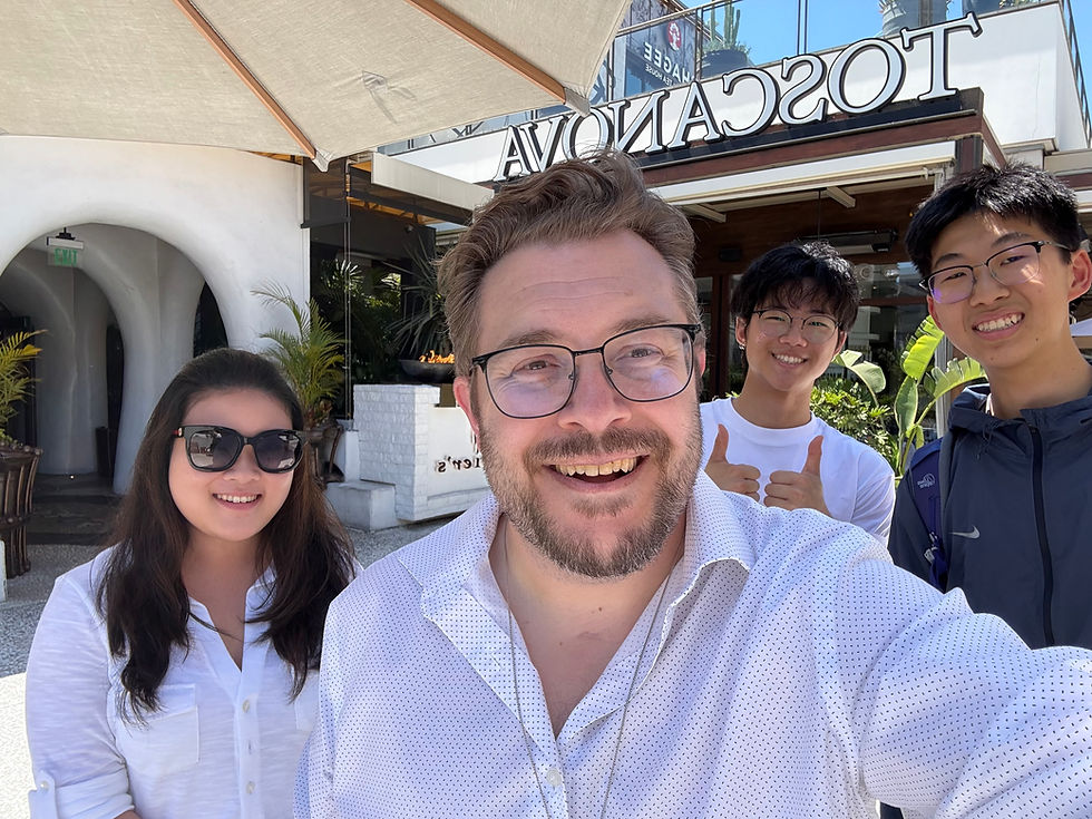 Four people smiling outside a restaurant with the sign "TOSCANO" visible. One person gives a thumbs-up. Sunny day, relaxed mood.