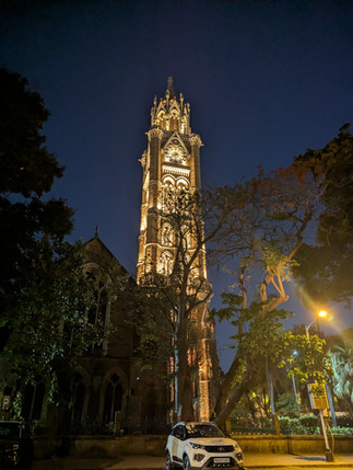 Illuminated Gothic clock tower rises above trees at night, with a white SUV parked below and a warm streetlight nearby.