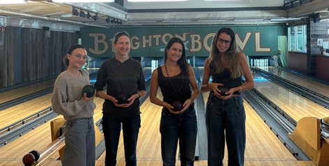 Four people holding bowling balls in a bowling alley. Visible text: Brighten Bowl.
