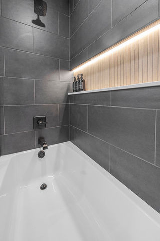 White bathtub, grey tiled shower, illuminated recess shelf with bottles.