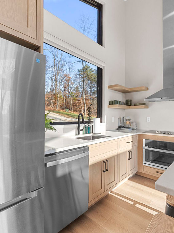 Modern kitchen with stainless steel appliances and large window overlooking woods.
