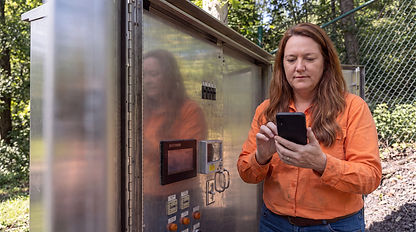 Technician in orange shirt using mobile phone near control box.