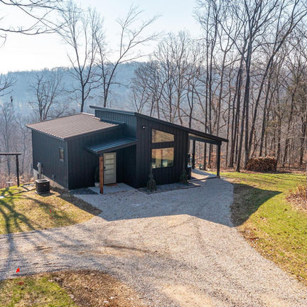 Modern dark house, angled roof, gravel driveway, on a wooded hillside.