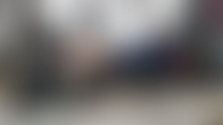 Man in navy outfit lounging on a lawn mower in a gravel yard, feet up, with trees and a fence in the background. Playful mood.