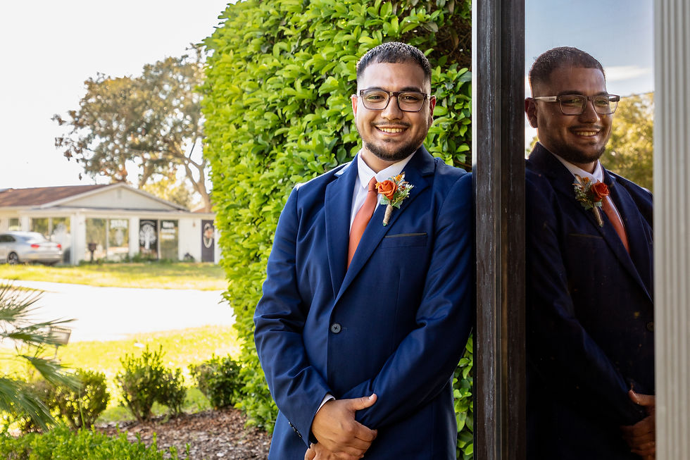 Man in a blue suit with orange tie and boutonniere smiles, leaning against a reflective surface. Green hedge and house visible in background.