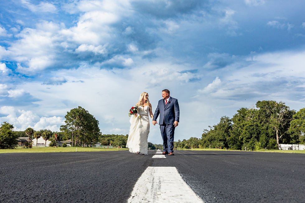 Bride and groom walk hand in hand on a rural road, surrounded by trees and under a cloudy blue sky. She holds a bouquet, both smiling.