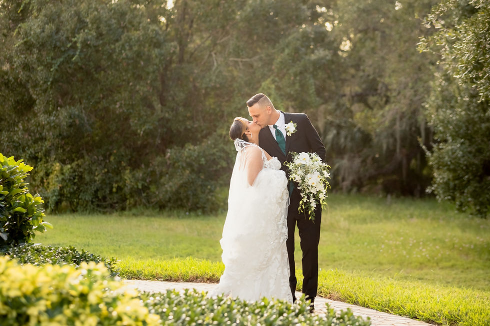 Bride and groom kiss, surrounded by lush greenery. She holds a white bouquet. Both are dressed formally, creating a romantic scene.