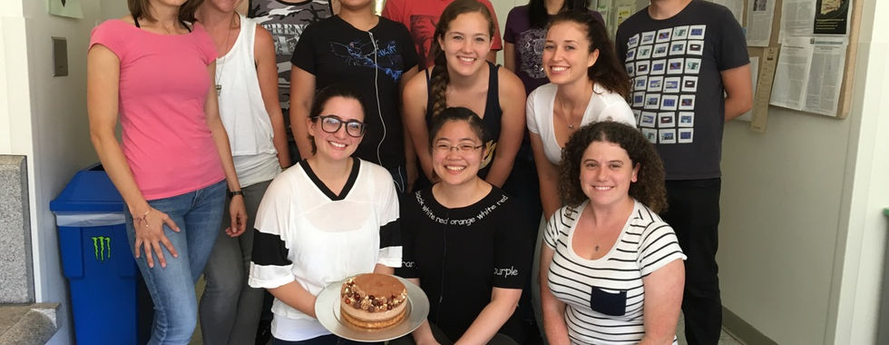 The team smiles as they pose in a UCLA hallway. Sofia, kneeling in front, holds the birthday cake
