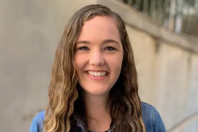Welcome to lab assistant Elizabeth, with long, wavy blonde hair, almond-shaped eyes, and a blue shirt, smiling in front of a wall.