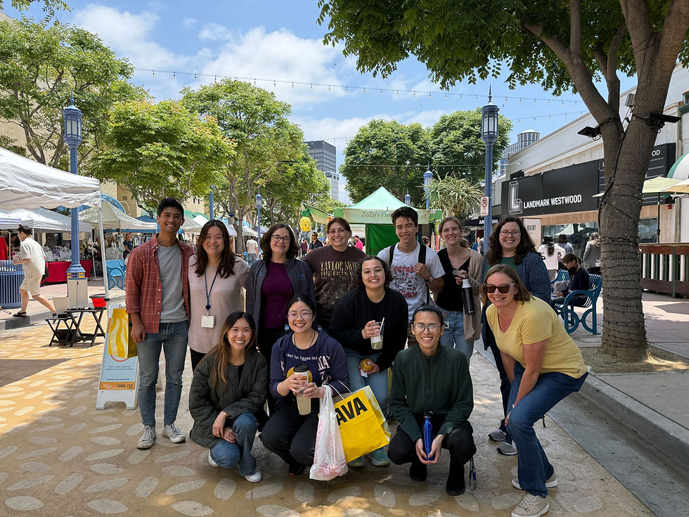 Members of the ETV lab smile for a group photo outdoors on a sunny day. They wear casual clothes and hold bags, bottles, and drink cups. In the background, people walk by, tents are set up, and many trees stand under a blue sky with few clouds.