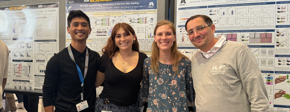 From right to left, Dr. Thomas, with slicked-back hair, clear-frame glasses, and a light sweatshirt with lab logos; Dr. Heidi, with loose blonde hair, a pendant necklace, and a floral blouse; Gabriella, with long auburn hair and bangs, wearing a patterned skirt and black blouse; and Dr. Alvin, with short styled hair, dark clothes, and a brown belt. All smile in front of their displayed posters.