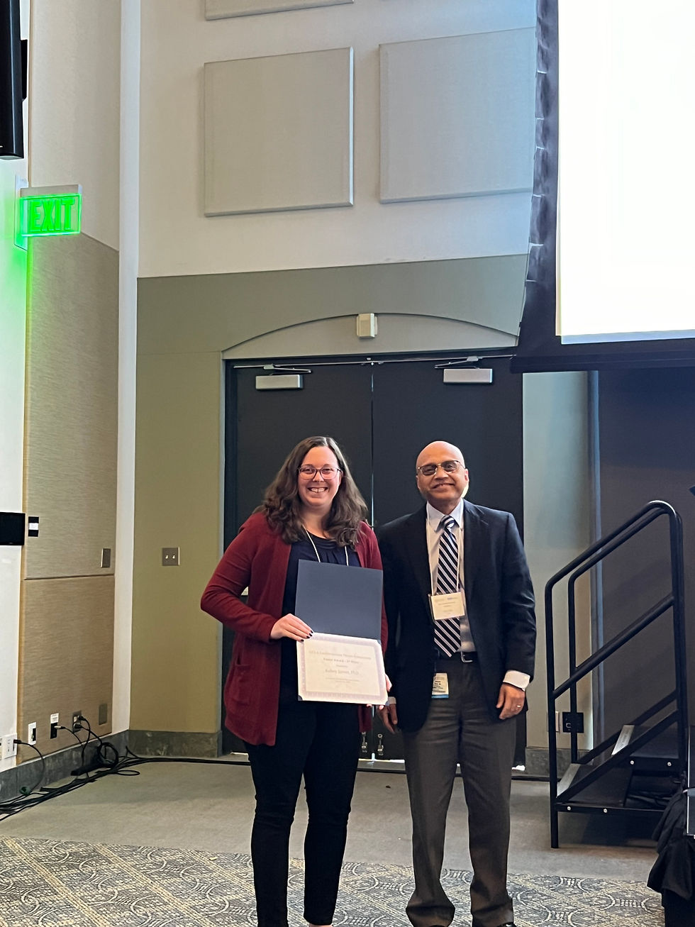 Kelsey, with loose brown hair and wearing dark clothing and a burgundy coat, smiles while holding a certificate presented by the man standing next to her, who is also smiling, bald, wearing glasses, and dressed in a suit jacket, tie, and dress pants.