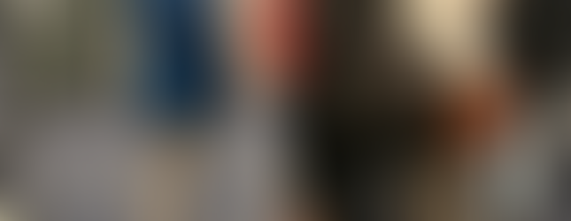 Kelsey, with shoulder-length wavy brown hair, wearing a blue coat, light-colored pants, and black shoes, is seen from behind, focused on a group of researchers lined up to hear her explain her poster, which is displayed on a board in a hall with other research presentations.