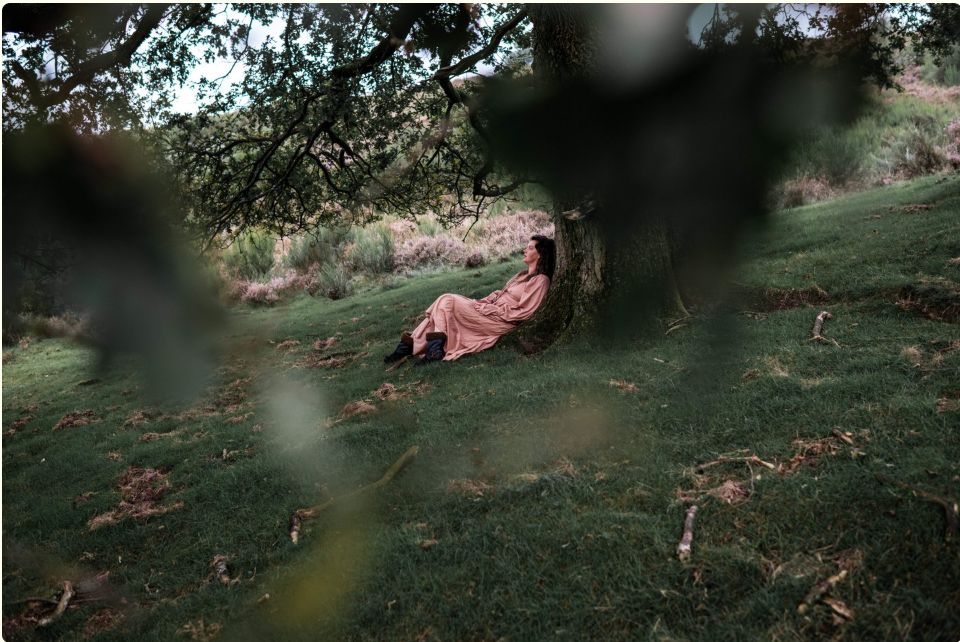 Vrouw in roze jurk leunend tegen een boom in een groen landschap, liggend breedbeeld (wide landscape)