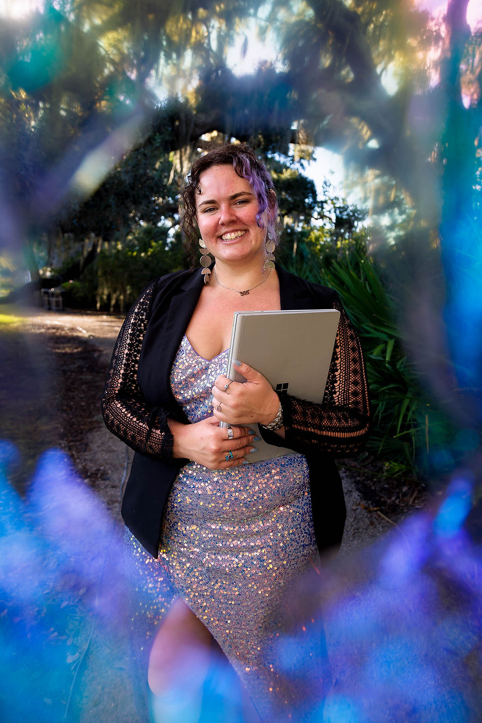 Smiling woman in a shimmering dress holds a laptop, framed by colorful light reflections in a lush, outdoor setting.