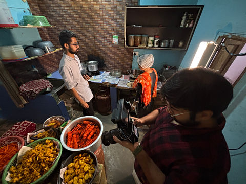 Behind-the-scenes shot of filmmakers documenting a micro-business food kitchen in Delhi, with trays of snacks in the foreground and the cook preparing dishes.