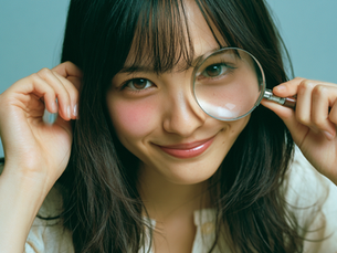 A smiling young woman with dark hair holding a magnifying glass over her eye to symbolize research, search engine optimization, and AI discovery