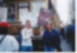 Protest scene with John and his friend holding anti-war signs, "WHAT WOULD JESUS DO?" and "NO BLOOD FOR OIL." Urban street and crowd in the background.