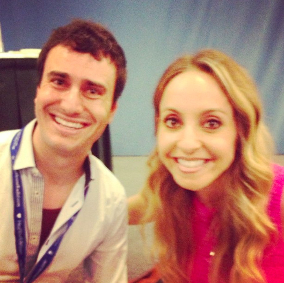 John Mothershead (noticably younger) and Gabrielle Bernstein smiling, John wearing a blue lanyard, Gabrielle in a pink top. Blurred blue background, bright and cheerful mood.