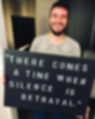 John smiling, holding a black sign with "There comes a time when silence is betrayal." written in stencil font. Indoor setting.