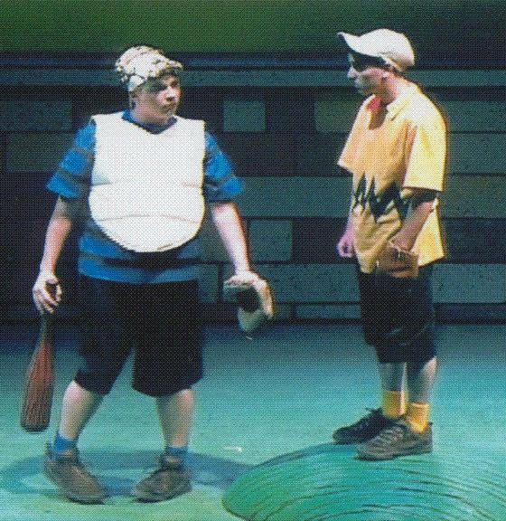 Two people in cartoon-like costumes stand on stage. One wears a yellow shirt with a zigzag pattern, the other a striped shirt with padding.