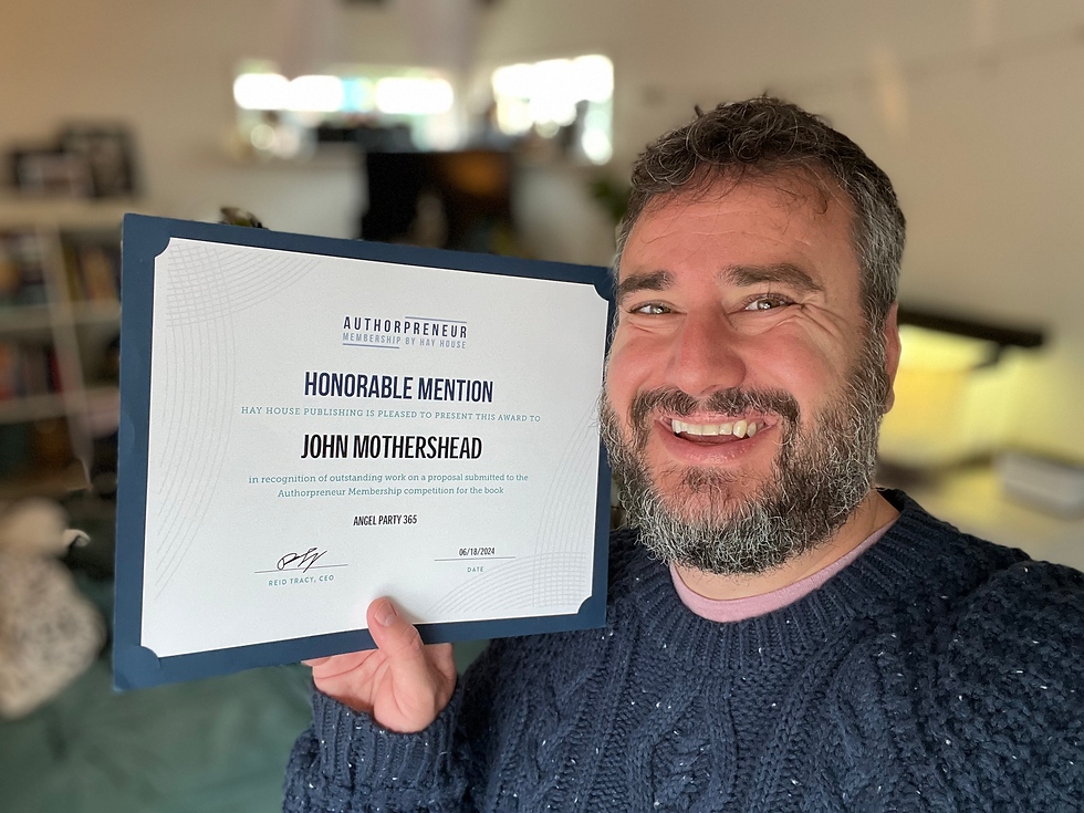 Smiling man holding a certificate with "Honorable Mention" text. Wearing a navy sweater in a cozy room with blurred shelves in the background.
