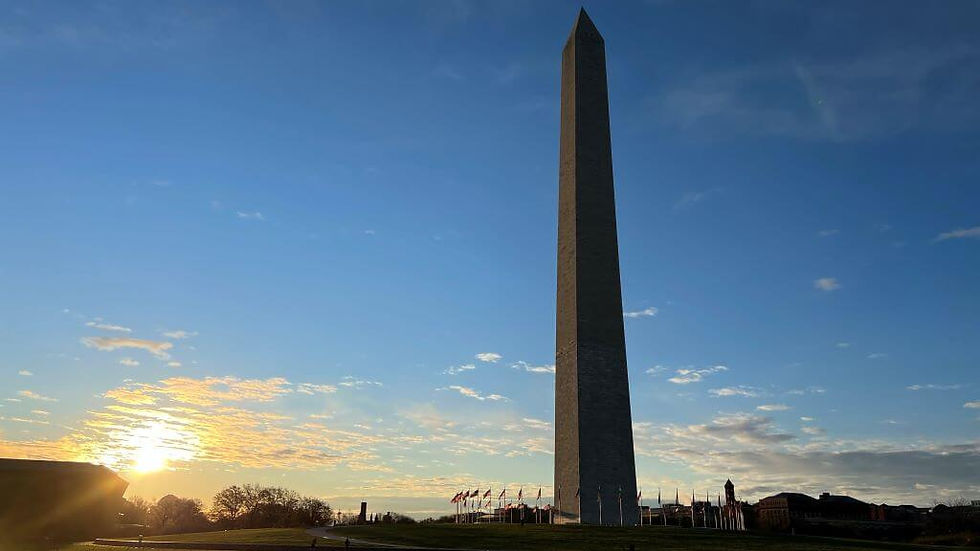 Tall obelisk against a blue sky at sunrise, surrounded by flags. A path and grass in the foreground create a peaceful, morning scene.